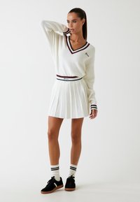 Witte geribde trui met V-hals en bordeaux details, gecombineerd met een geplooide witte rok. Zwarte sneakers en gestreepte sokken maken de look compleet.