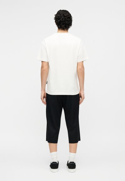 Άνδρας με σγουρά μαλλιά, που φοράει απλό λευκό T-shirt, μαύρο κοντό παντελόνι, λευκές κάλτσες και μαύρα αθλητικά παπούτσια, στέκεται με την πλάτη προς τον θεατή.