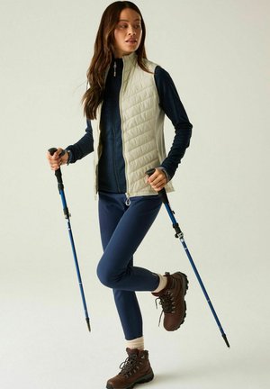 Woman wearing a light vest, navy jacket, and hiking boots holding blue trekking poles, standing on one leg against a plain background.