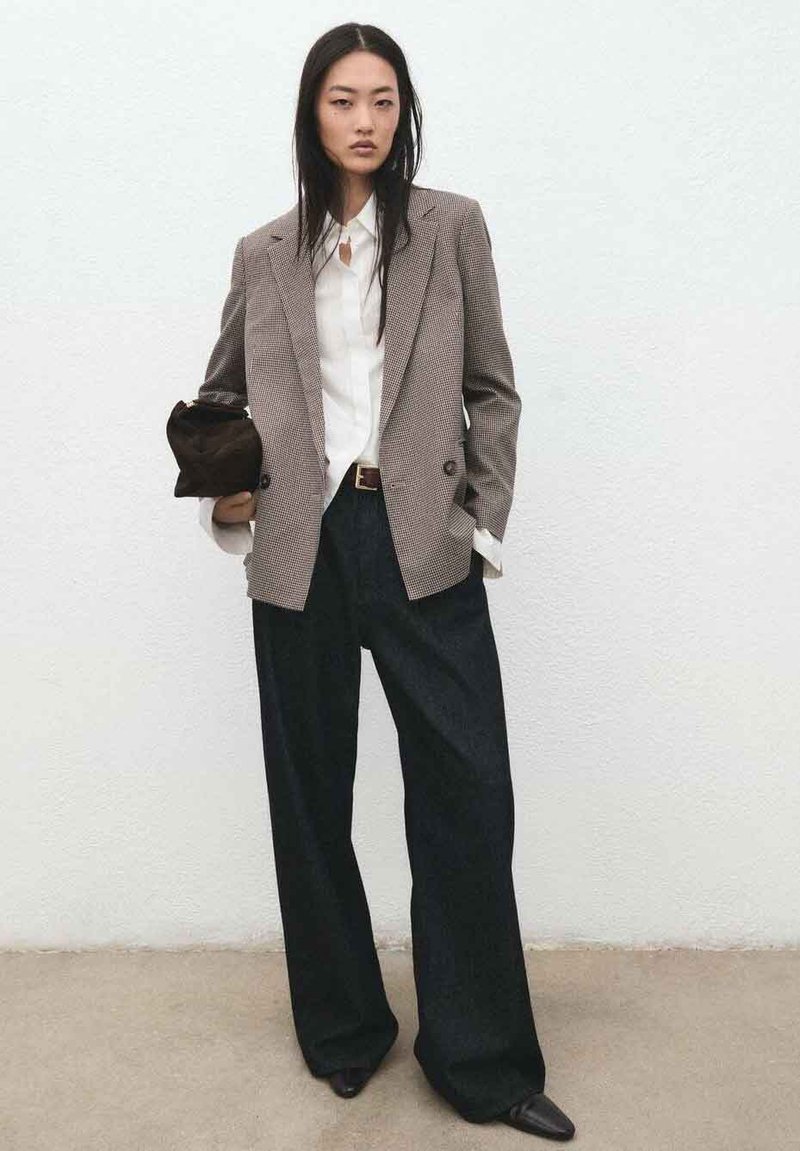 Bruine houndstooth blazer over een wit overhemd, donkergekleurde jeans met hoge taille, en een bruine clutch. De outfit wordt gecompleteerd met zwart puntige schoenen.