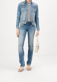 Lichtblauwe denimjack met twee borstzakken, gecombineerd met een gestreept shirt en vervaagde blauwe jeans. Beige schoudertas en tan flip-flops.