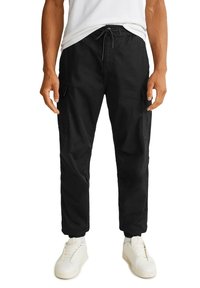 Homme portant un pantalon cargo noir avec des poignets élastiques, une taille à cordon et des baskets blanches, debout les mains détendues le long du corps.