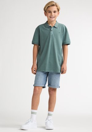 Polo vert à col, associé à un short en denim clair. Des baskets blanches et des chaussettes rayées complètent cette tenue décontractée.