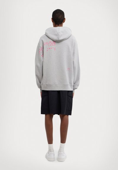Sweat-shirt à capuche gris avec une coupe décontractée, arborant un texte graphique rose au dos. Associé à un short noir et des baskets blanches.