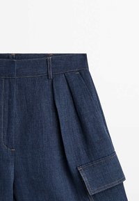 Pantalon en denim bleu foncé avec plis, large ceinture avec passants et poche à rabat sur le côté sur fond blanc uni.