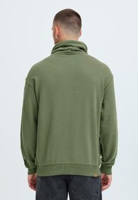 Pullover verde con colletto alto, maniche lunghe, polsini e orlo a coste. Presenta un tessuto texture e un piccolo etichetta di marca nella parte inferiore.