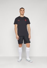 T-shirt sportivo nero con logo rosso, abbinato a pantaloni corti neri. Il modello indossa calzini bianchi fino al ginocchio e scarpe da calcio argento con dettagli rosa.