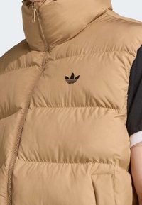 Béžová prošívaná vesta s vysokým límcem, předním zipem a černým logem Adidas na hrudi. Má měkkou, hladkou texturu a boční kapsu.