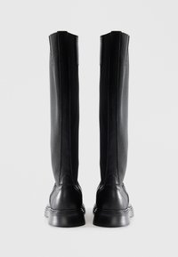 Une paire de bottes noires en cuir hautes avec des panneaux en tissu texturé, vue de l'arrière sur un fond clair.