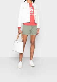 Giacca di denim bianca sopra una felpa rossa con stampa "A", pantaloncini verdi, sneaker bianche e borsa bianca con manico intrecciato.