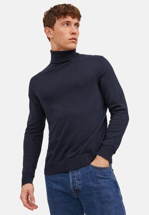 Nuori mies, jolla on yllään laivastonsininen pooloneule ja siniset farkut, poseeraa yhdellä kädellä taskussa ja katsoo sivulle yksinkertaisella taustalla.