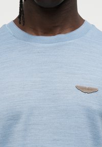 Ανοιχτό μπλε βαμβακερό T-shirt με υφή υφάσματος, ριμπ ύφασμα στη λαιμόκοψη και καφέ ταμπελάκι με το λογότυπο της Aston Martin στο αριστερό στήθος.