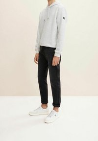 Ljust grå huvtröja med ett texturerat mönster, svarta jeans och vita sneakers. Huvtröjan har dragsnören och en liten logotyp på ärmen.