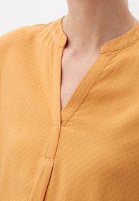 Blouse jaune moutarde avec un col en V, un motif texturé et des manches courtes. Le tissu semble léger et doux.