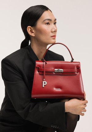 Femme aux cheveux foncés portant un blazer noir tenant un sac à main rouge brillant avec une anse supérieure et un fermoir argenté devant un fond uni.