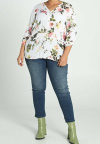 Femme portant un chemisier blanc à motifs floraux avec manches trois-quarts, jean skinny bleu et bottines à talons vert clair, debout devant un fond blanc.