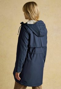 Námornícky modrý nepremokavý parka s textúrovanou, fleece podšívkou na kapucni, nastaviteľnými šnúrkami a ventilačným otvorom na chrbte.