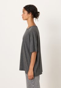 Siv velika majica s kratkimi rokavi in širokim izrezom, narejena iz mehke tkanine. Model nosi svetlo sive, visokopričesane kavbojke.