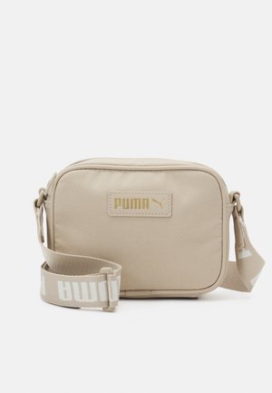 Beżowa prostokątna torba Puma na ramię z regulowanym paskiem i złotą naszywką z logo na przedniej stronie.