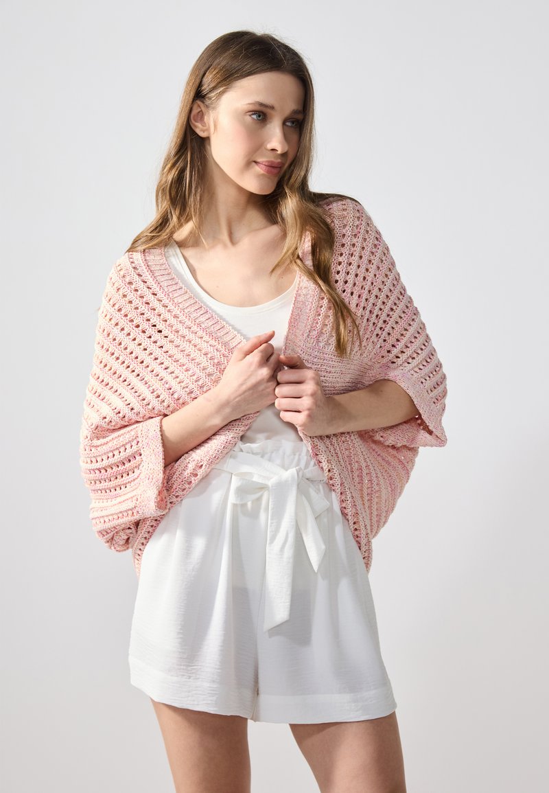 Lätt, stickad sjal i gradient rosa. Öppen väv med vida ärmar. Modellen bär den över en vit tanktopp och högmidjade shorts.