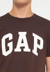 Hnědý bavlněný tričko s velkým bílým logem "GAP" v tučném, sportovním písmu. Klasický kulatý výstřih a krátké rukávy.