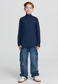 Granatowa koszulka z długim rękawem i golfem zestawiona z jasnoniebieskimi, luźnymi dżinsami cargo z bocznymi kieszeniami oraz białymi trampkami z ciemnymi akcentami.