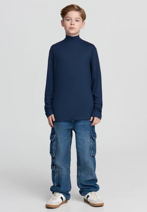 Granatowa koszulka z długim rękawem i golfem zestawiona z jasnoniebieskimi, luźnymi dżinsami cargo z bocznymi kieszeniami oraz białymi trampkami z ciemnymi akcentami.