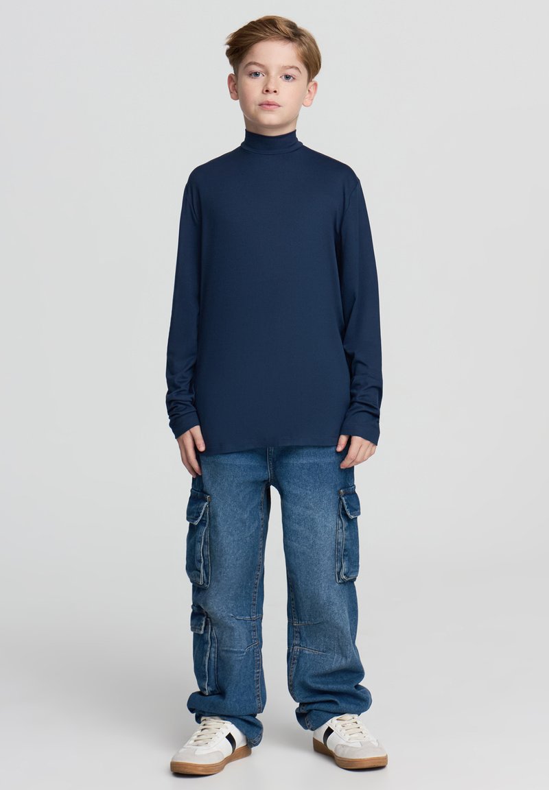 Granatowa koszulka z długim rękawem i golfem zestawiona z jasnoniebieskimi, luźnymi dżinsami cargo z bocznymi kieszeniami oraz białymi trampkami z ciemnymi akcentami.