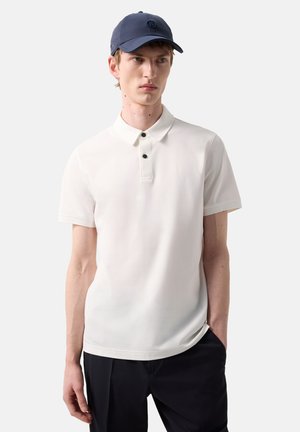 Young man wearing a white short-sleeve polo shirt with black buttons, black pants, and a dark blue baseball cap, standing with one hand in pocket.