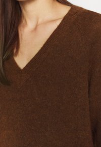 Pull marron à col en V en matière douce et texturée, avec une coupe décontractée et des coutures apparentes le long de l'encolure et du corps.