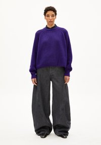 Übergroßer lila gestrickter Pullover mit Struktur, hohem Kragen und langen Ärmeln, kombiniert mit weit geschnittenen dunkelgrauen Jeans. Schwarze spitze Schuhe.