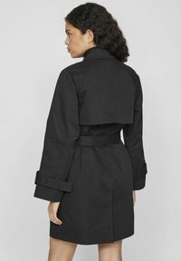 Svart trench coat med bälte i midjan, med långa ärmar, hög krage och ett lagerformat bakdesign. Slät, matt tygtextur.