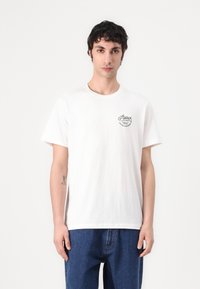 Weißes Baumwoll-T-Shirt mit Rundhalsausschnitt, kurzen Ärmeln und einem schwarzen Logo-Detail auf der linken Brust, kombiniert mit blauen Denim-Jeans.
