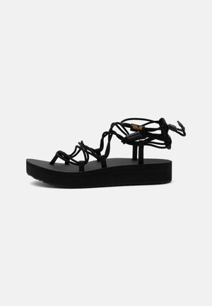 Sandalo platform nero con cinturini, suola testurizzata e chiusura regolabile con lacci alla caviglia, mostrato su sfondo bianco.