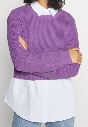 Person with crossed arms wearing a white collared shirt under a cropped purple knit sweater and light blue jeans.