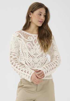 Femme aux longs cheveux portant des boucles d'oreilles créoles dorées et un pull en crochet blanc détaillé, pantalon beige, regardant sur le côté avec les mains jointes.