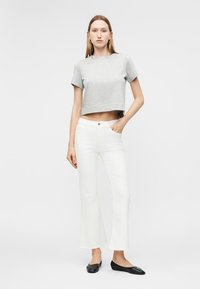 Frau steht mit der Hand in der Tasche, trägt ein graues, kurz geschnittenes T-Shirt, weiße, hoch taillierte Jeans und schwarze Ballerinas vor weißem Hintergrund.