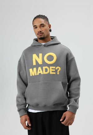Grijze hoodie met een voorzak, met grote gele tekst "NO MADE?" in een vet lettertype. Zacht materiaal met een iets losse pasvorm.