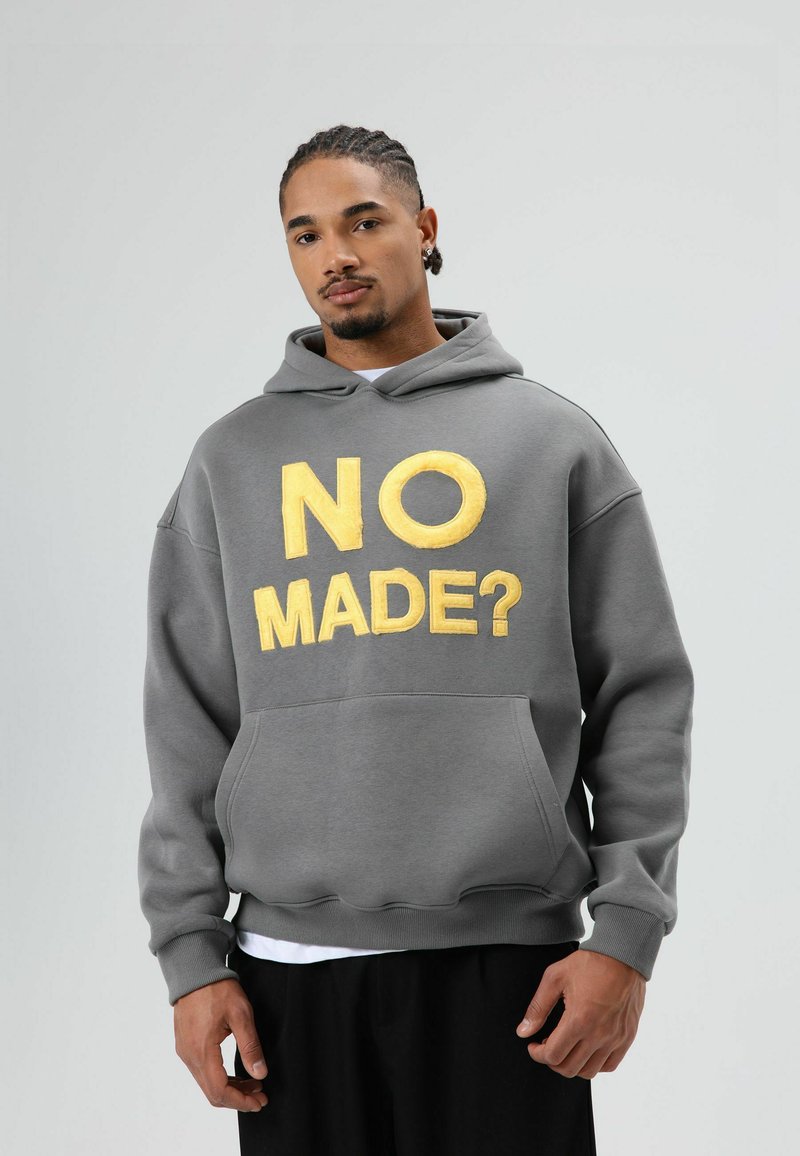 Grijze hoodie met een voorzak, met grote gele tekst "NO MADE?" in een vet lettertype. Zacht materiaal met een iets losse pasvorm.