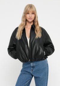 Blonde Frau trägt eine schwarze Lederbomberjacke über einem weißen Oberteil und blauen Jeans und steht mit den Händen in den Taschen der Jacke.