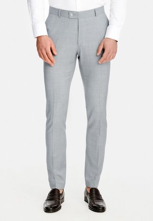 Mężczyzna ubrany w dopasowane, jasnoszare spodnie wizytowe, białą koszulę i czarne skórzane mokasyny, stojący z rękami swobodnie opuszczonymi wzdłuż ciała.