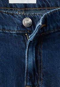 Primer plano de unos vaqueros de mezclilla azul oscuro, que presentan un botón metálico, cremallera y costuras marrones en contraste. Textura suave y diseño clásico.