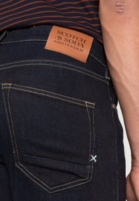 Mörkblå denimjeans med en beige läderetikett som har "Scotch & Soda"-logotypen. Anmärkningsvärda blå och vita sömnadsdetaljer nära fickan.