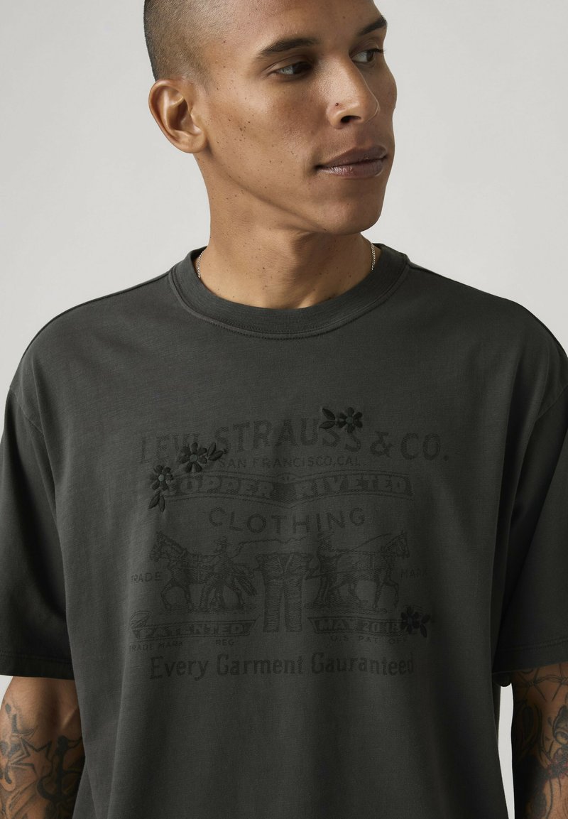 Homme à la tête rasée, portant un t-shirt Levi Strauss & Co. sombre et oversize avec une broderie florale et des tatouages visibles sur les avant-bras.