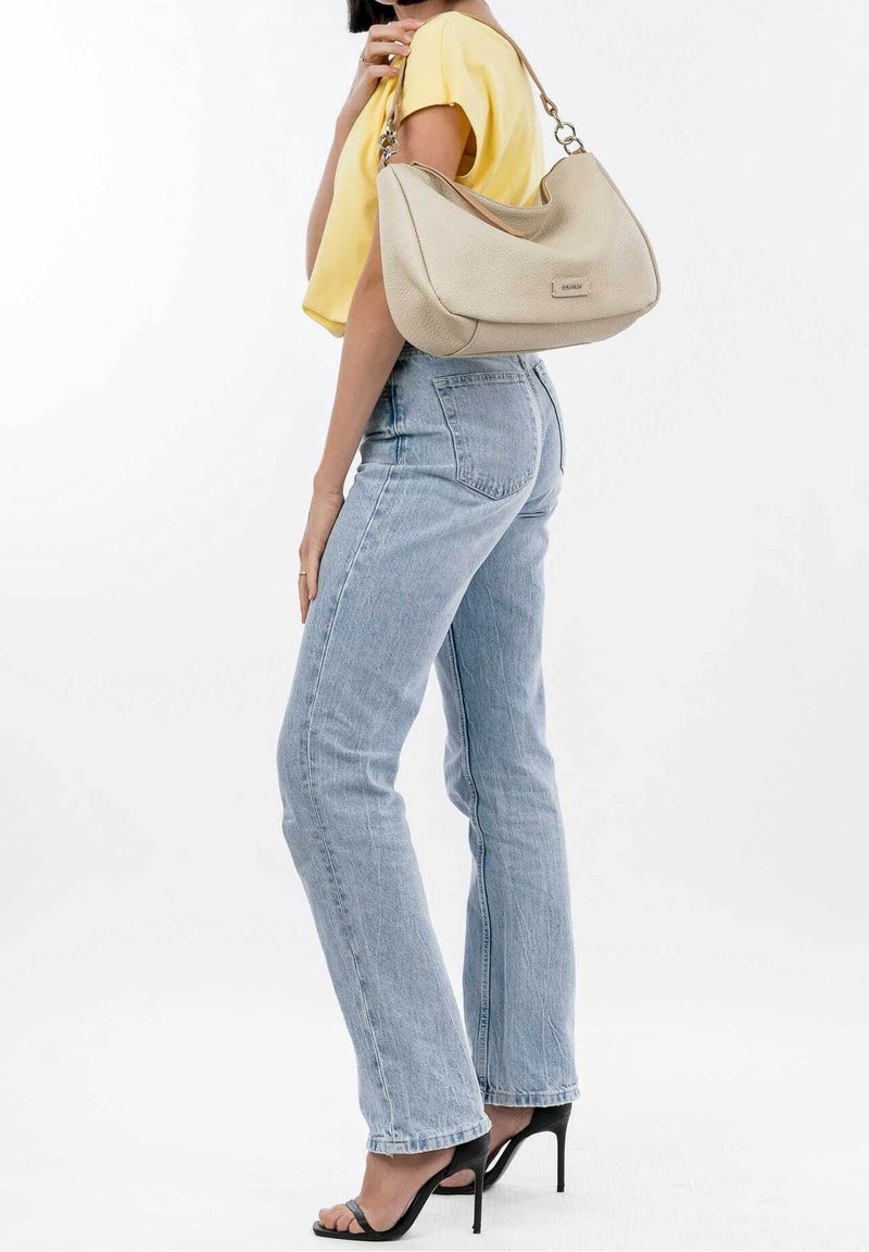 Femme en jean bleu clair et haut jaune tenant un sac à épaule beige, portant des sandales noires à talons hauts, posant sur fond blanc.