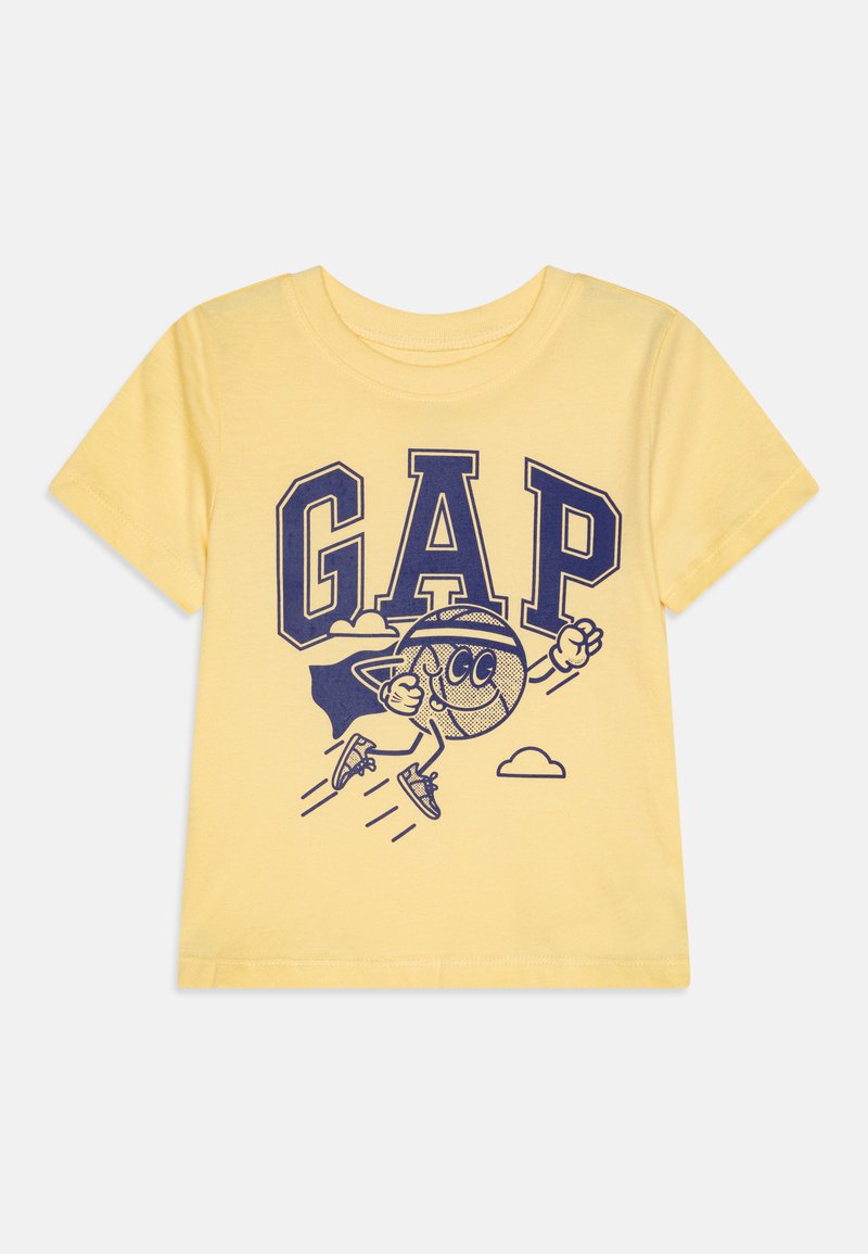 Gul t-skjorte i bomull med korte ermer, med en stor grafikk av "GAP" i marineblått og en tegneseriefigur i kappe som flyr blant skyer.