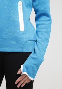 Blå långärmad träningströja med vit dragkedjeficka och tumhål på ärmarna, som bärs över svarta leggings mot en grå bakgrund.