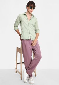 Camisa verde claro de botones con patrón texturizado, combinada con pantalones sueltos rosa y zapatillas blancas, exhibida sobre un taburete de madera.
