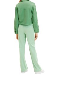 Giacca verde cropped con orlo sfilacciato, abbinata a pantaloni a zampa d'elefante verde chiaro e sneakers bianche, caratterizzata da una texture liscia e un design aderente.