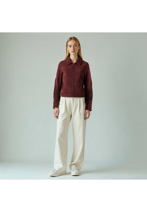 Bordó színű velúr dzseki rojtos részletekkel az ujjakon, cipzáras záródással és hegyes gallérral; világos krémszínű nadrággal és fehér tornacipővel párosítva.